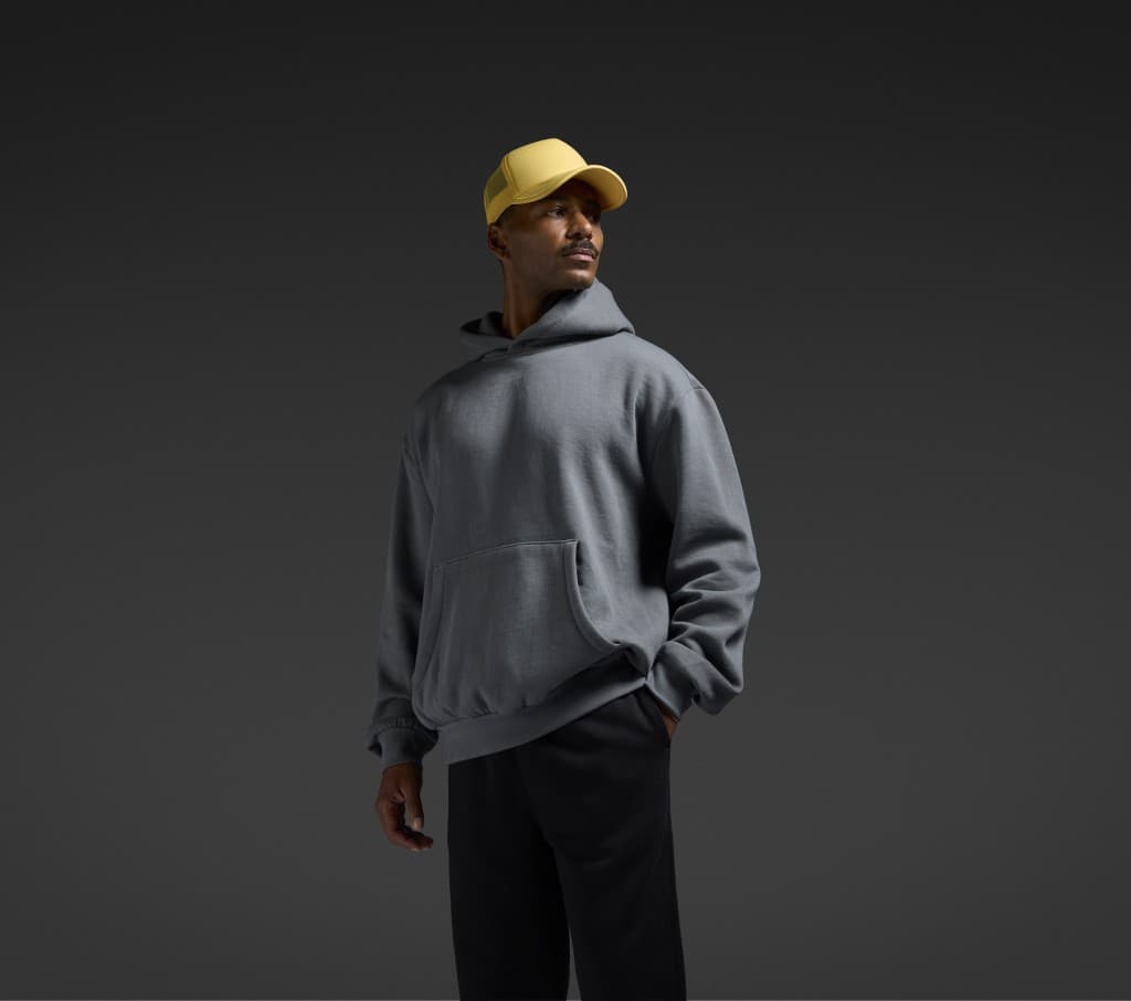 Model in a charcoal AS Colour hood and a yellow trucker cap against a dark backdrop.
