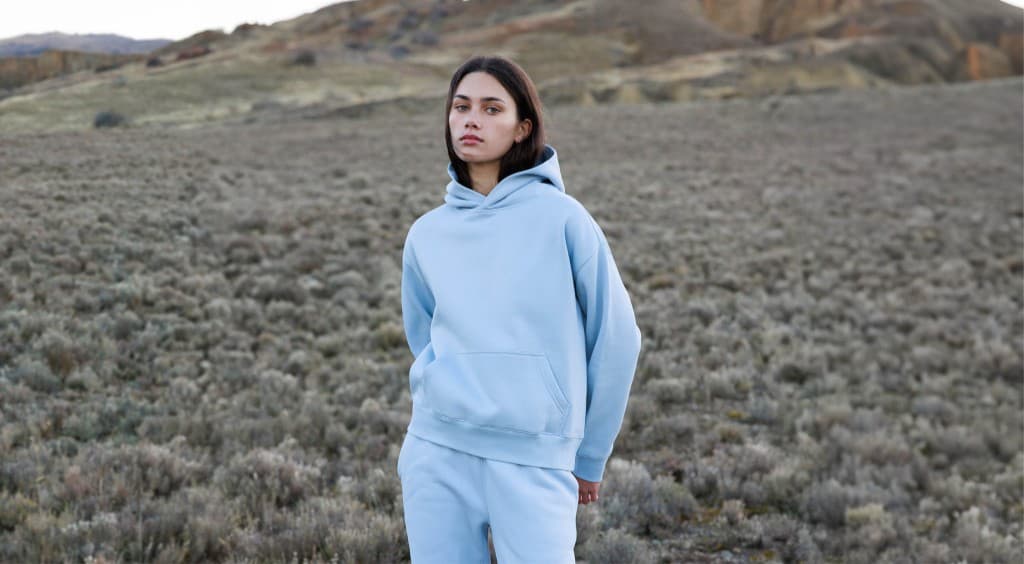 Model wearing a pale blue AS Colour hood and track pants in an open landscape.