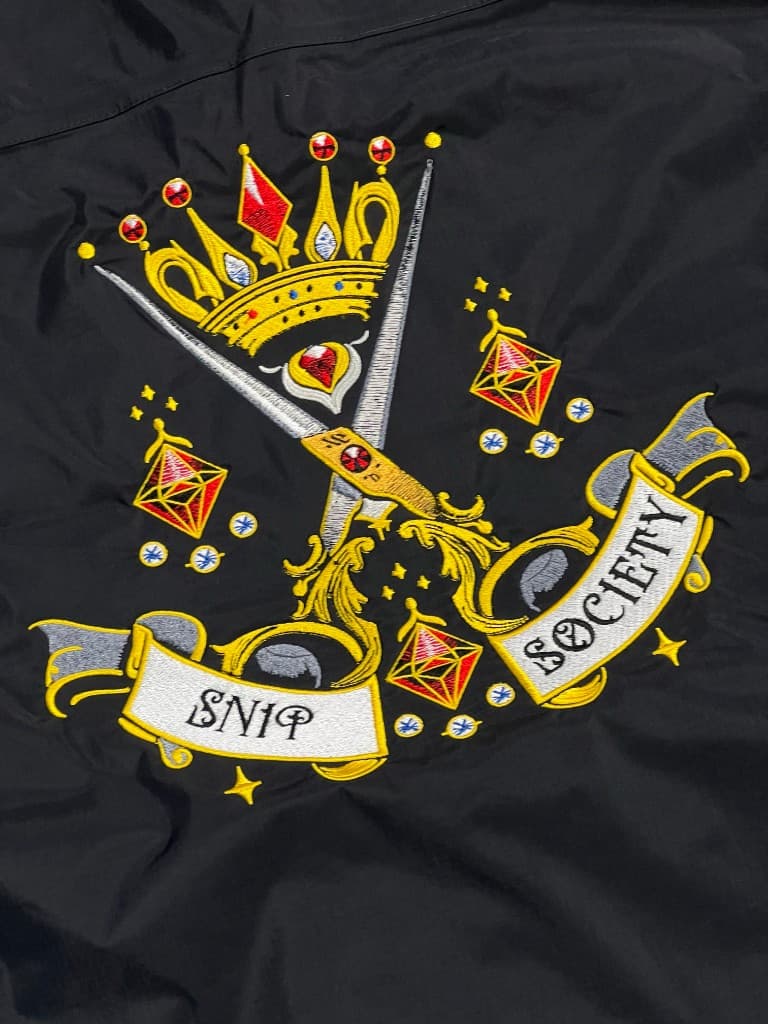 Detailed gold and silver embroidery on black fabric: crossed scissors, crown, gems, and Snip Society banner lettering.