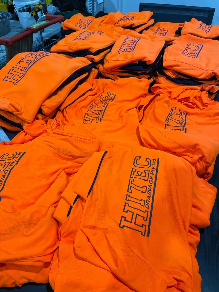Bulk stack of hi-vis orange workwear with navy screen-printed Hitec Drainage branding.